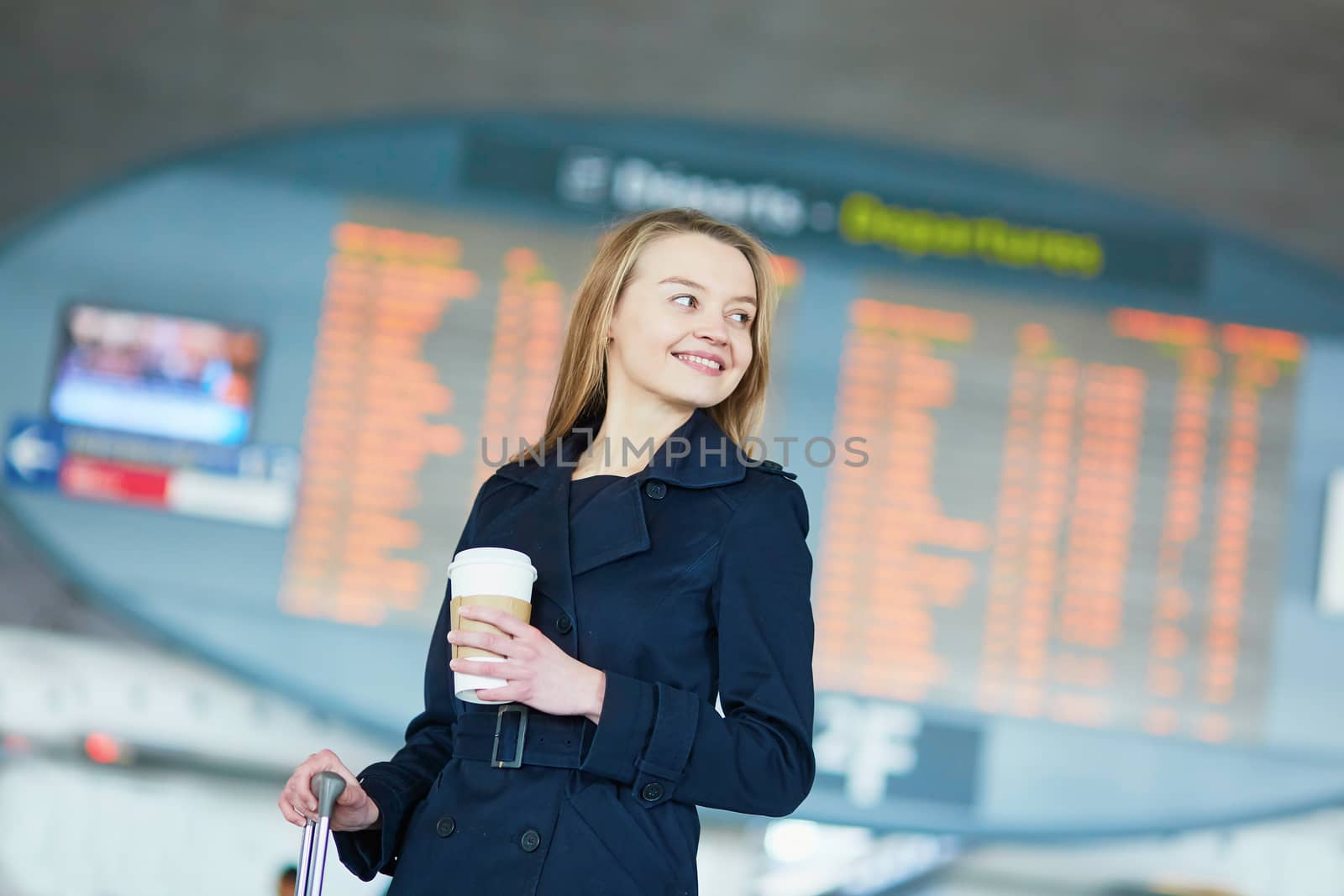 Young woman in international airport near the flight information board, with hot beverage to go