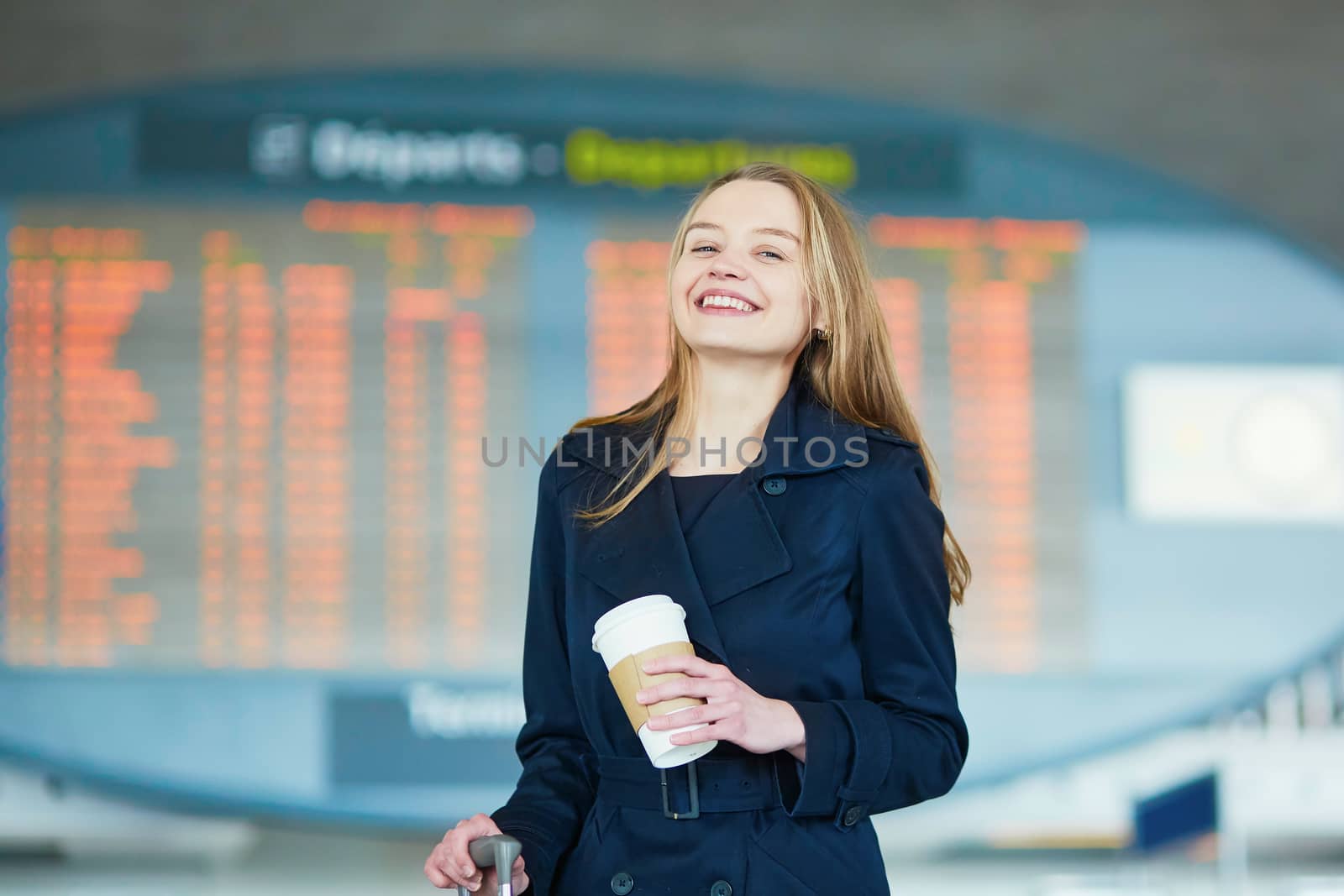 Young woman in international airport near the flight information board, with hot beverage to go