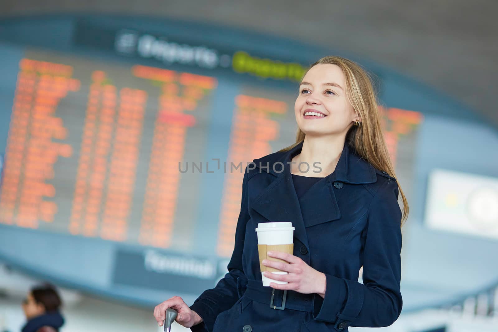 Young woman in international airport near the flight information board, with hot beverage to go