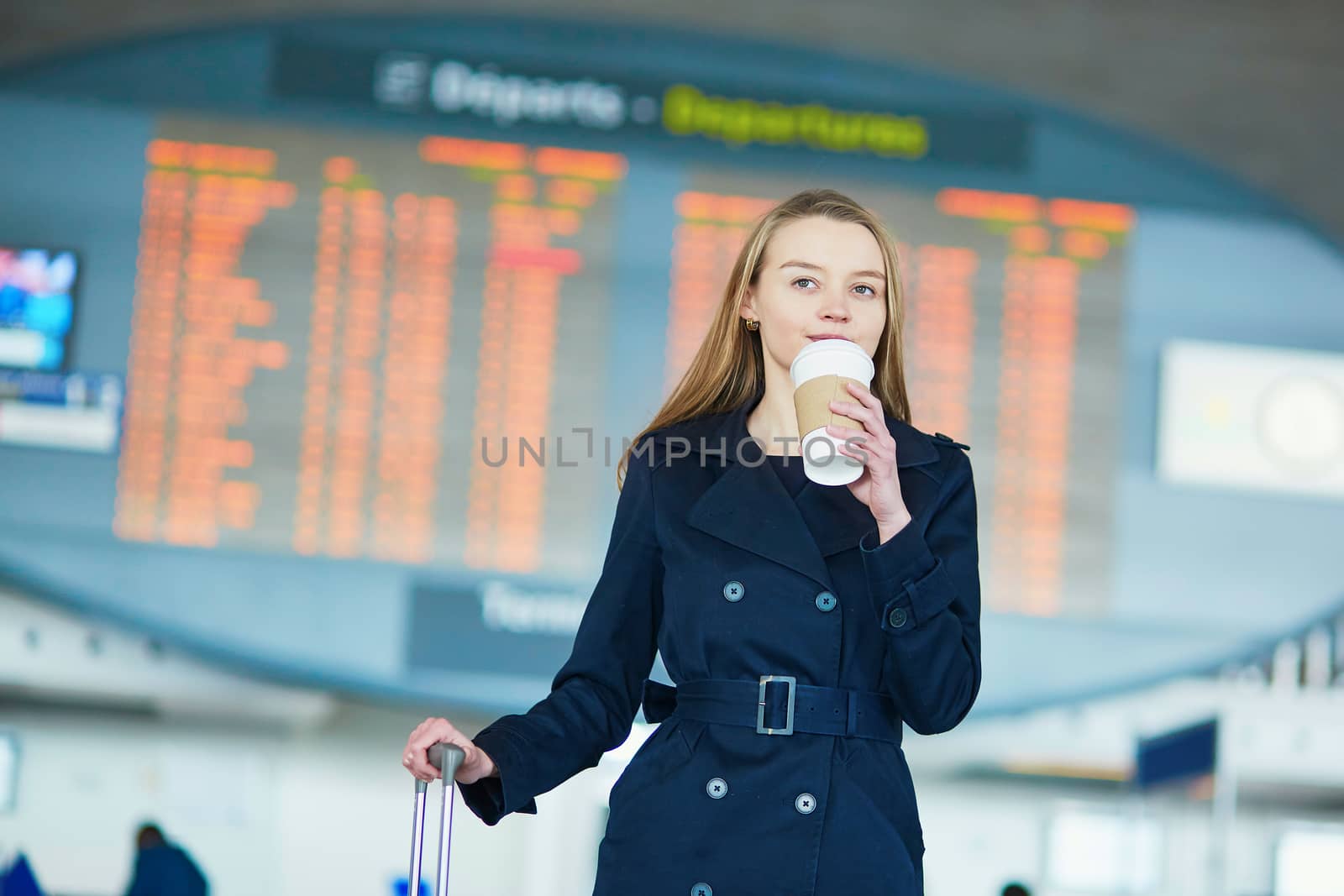 Young woman in international airport near the flight information board, with hot beverage to go