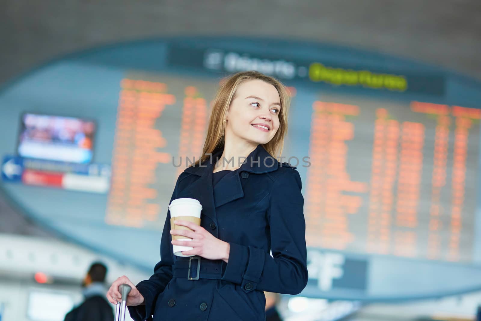 Young woman in international airport near the flight information board, with hot beverage to go