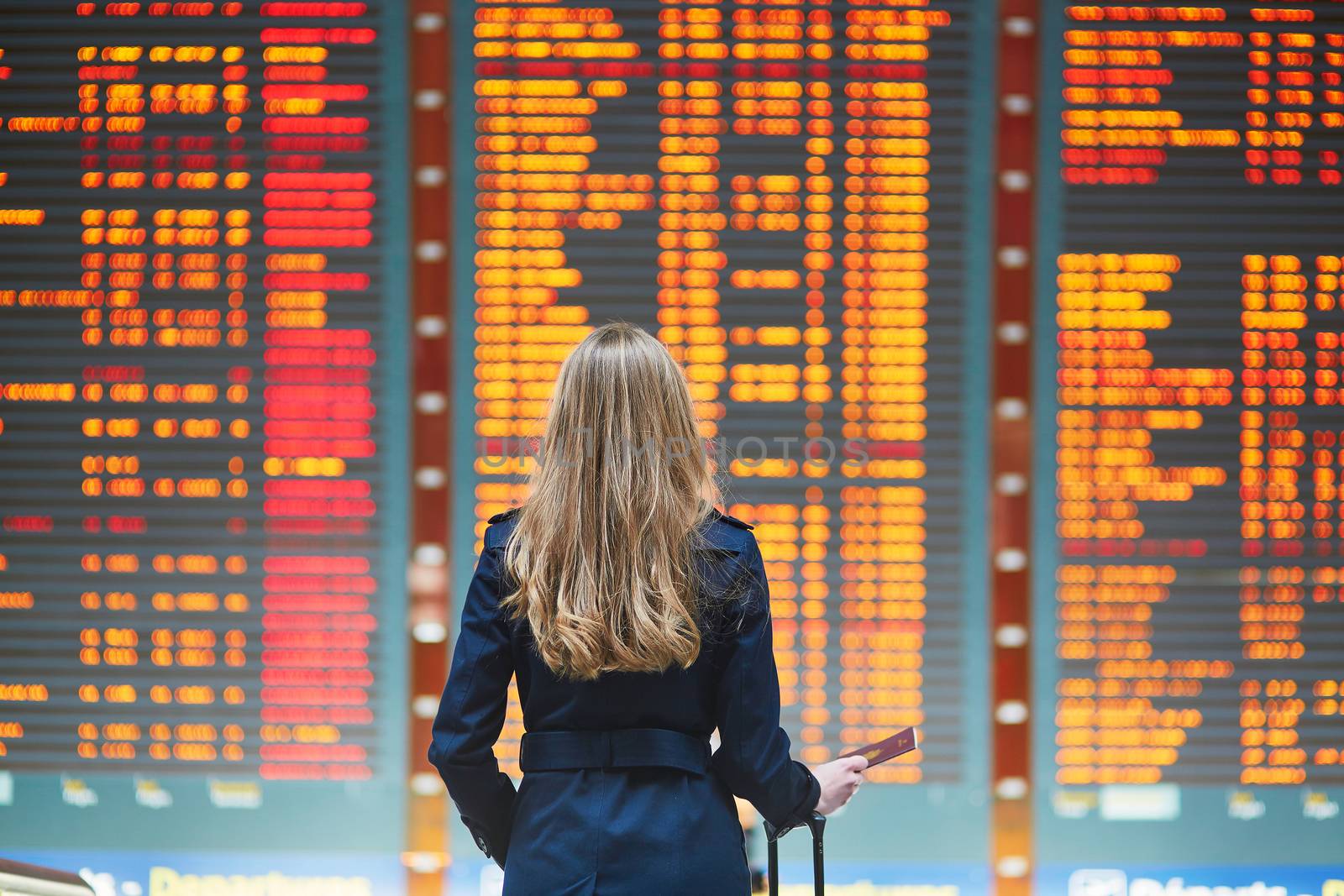 Young woman in international airport looking at the flight information board, holding passport in her hand, checking her flight