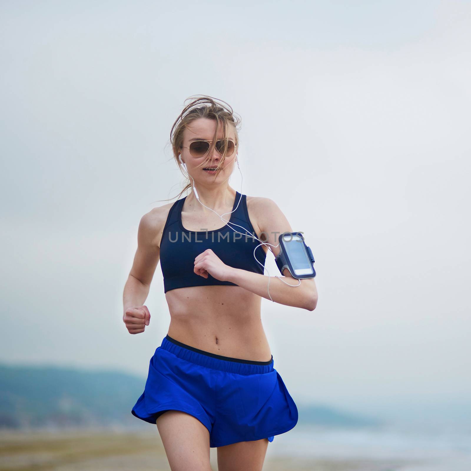 Running fitness woman wearing phone armband monitor tracker. Jogger running fast on beach and listening to music using earphones. Fitness and healthy lifestyle concept