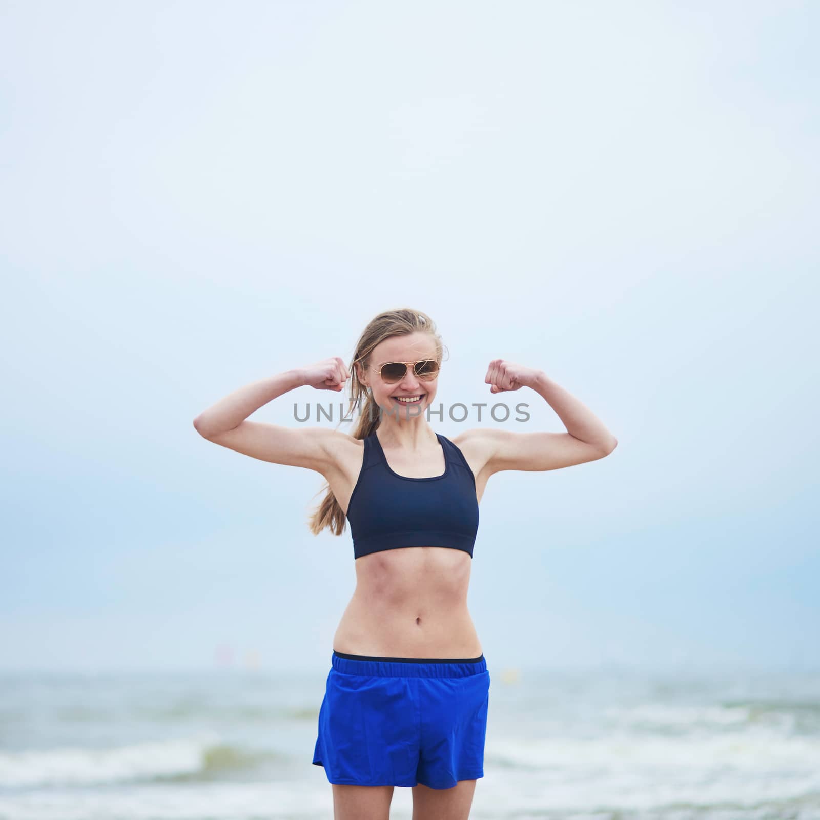 Healthy fitness runner girl wearing sunglasses showing her biceps. Young European woman on beach cardio training taking a rest during workout