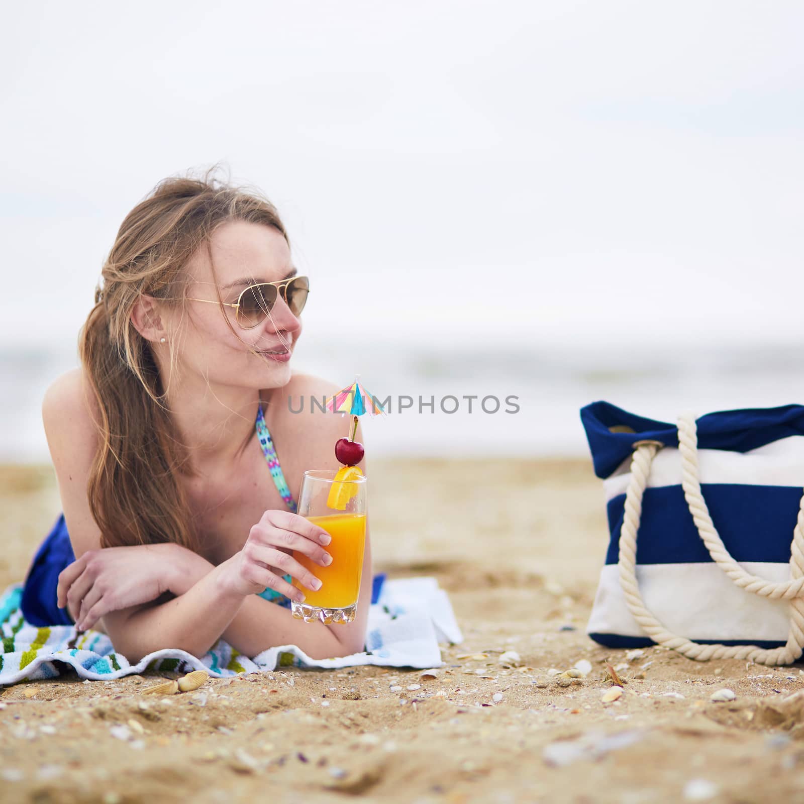 Beautiful young woman relaxing and sunbathing on beach, drinking delicious fruit or alcohol cocktail with paper umbrella, beach bag on sand near the model