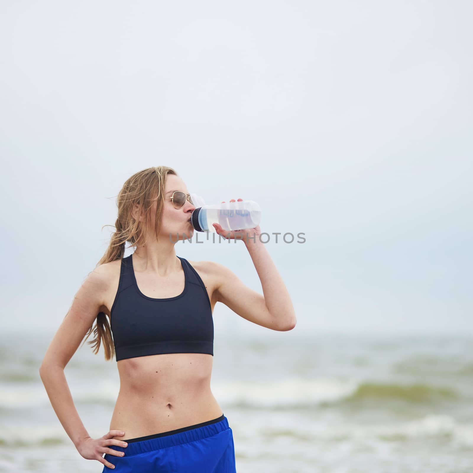 Healthy fitness runner girl drinking water from plastic bottle on running break. Young European woman on beach cardio training taking a rest during workout. Healthy lifestyle, sport and hydration concept