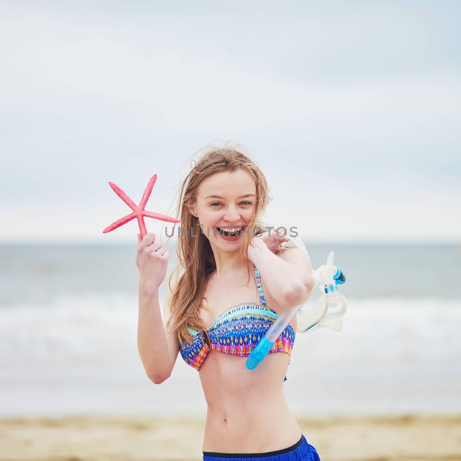 Happy young woman in bikini with snorkelling equipment and pink starfish enjoying summer vacation holidays by ocean or sea