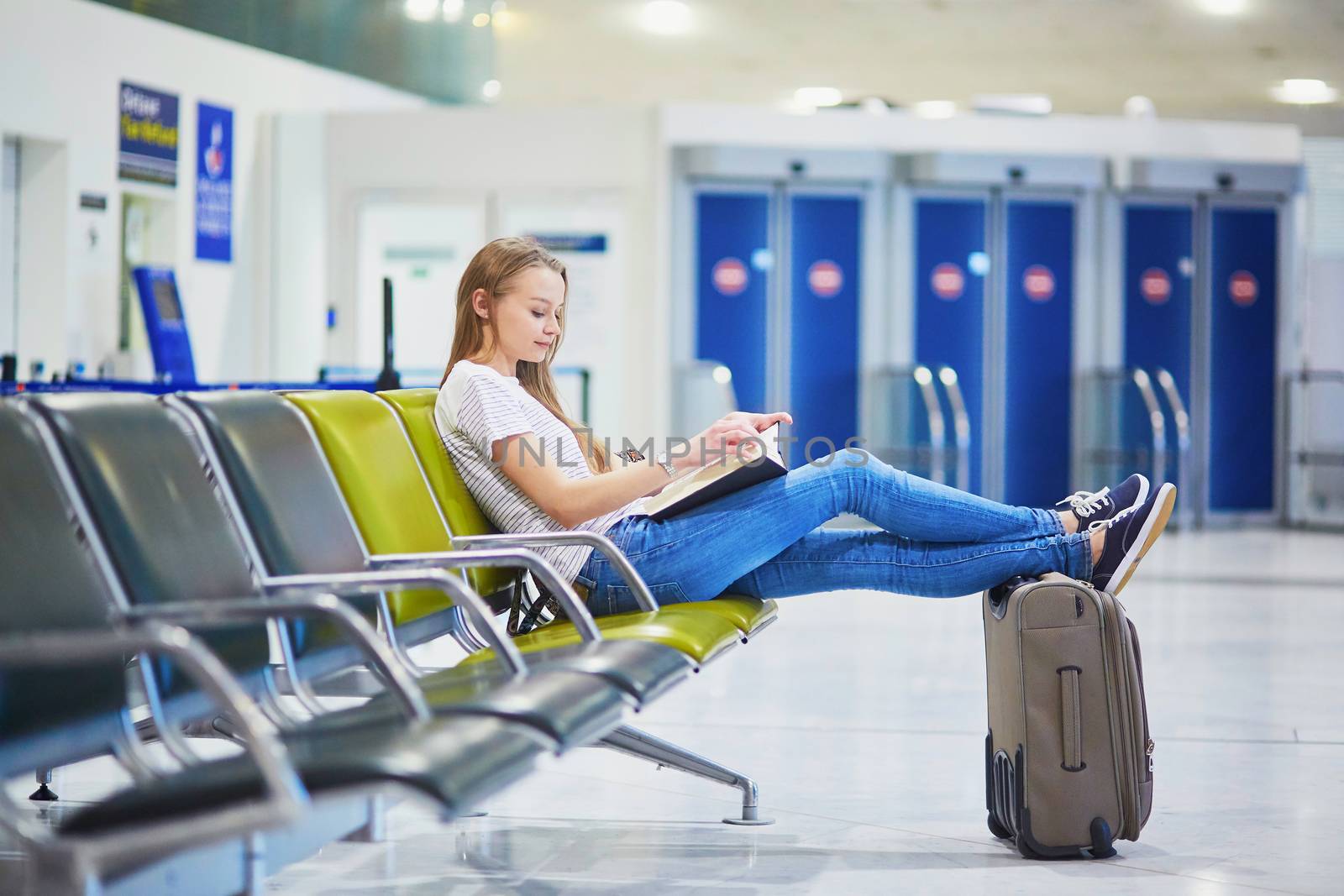 Young traveler with carry on luggage in international airport reading a book while waiting for her flight