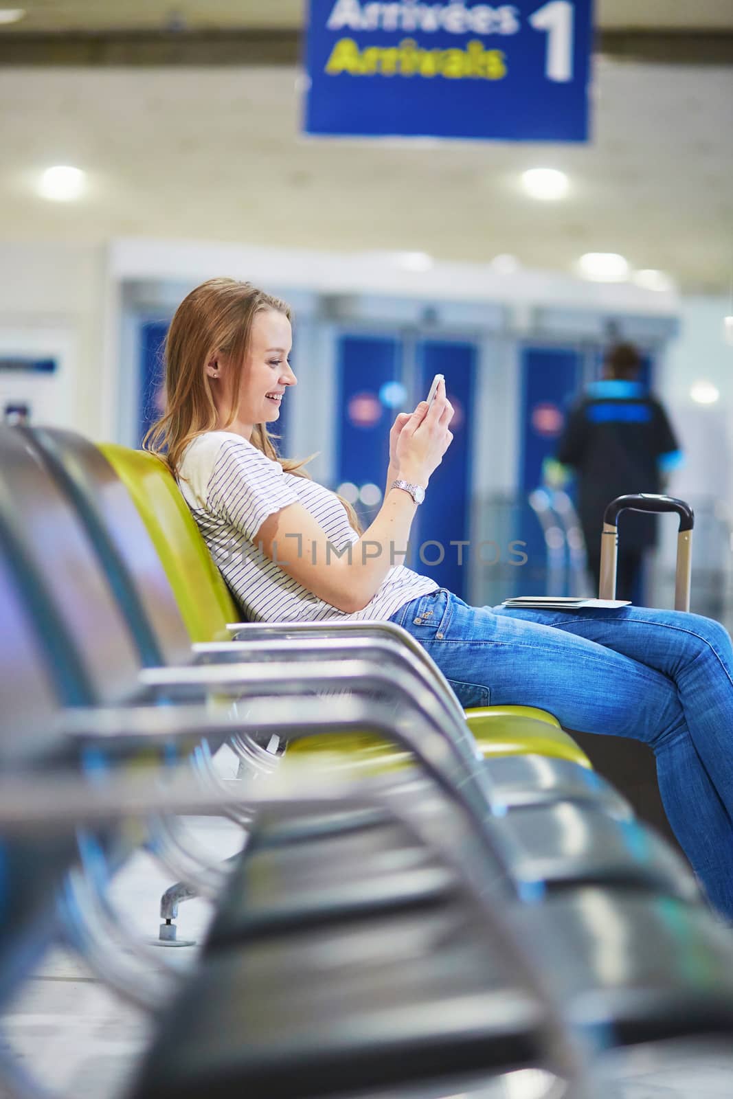 Beautiful young tourist girl with backpack and carry on luggage in international airport, checking her mobile phone while waiting for flight