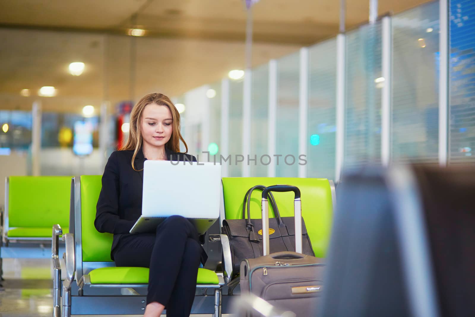 Young elegant business woman with hand luggage in international airport terminal, working on her laptop while waiting for flight