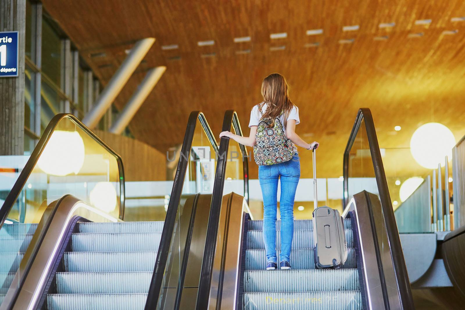 Beautiful young tourist girl with backpack and carry on luggage in international airport, on escalator