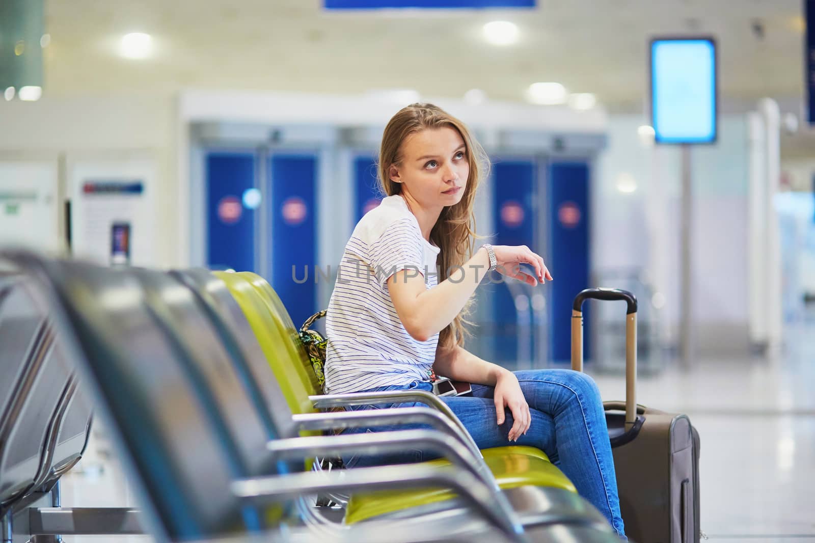 Beautiful young tourist girl with backpack and carry on luggage in international airport, waiting for her flight, looking upset. Delayed or canceled flight concept