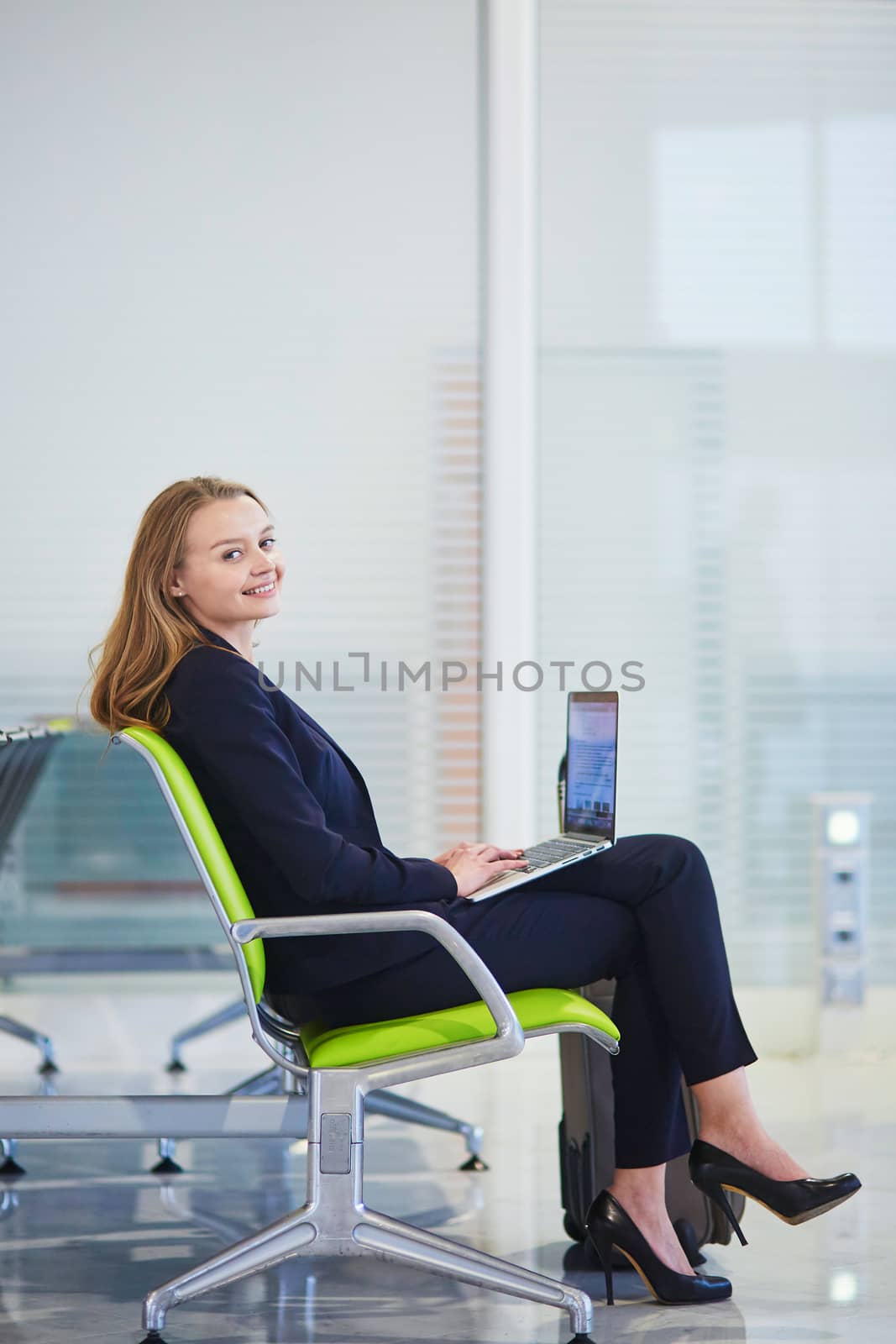 Young elegant business woman with hand luggage in international airport terminal, working on her laptop while waiting for flight