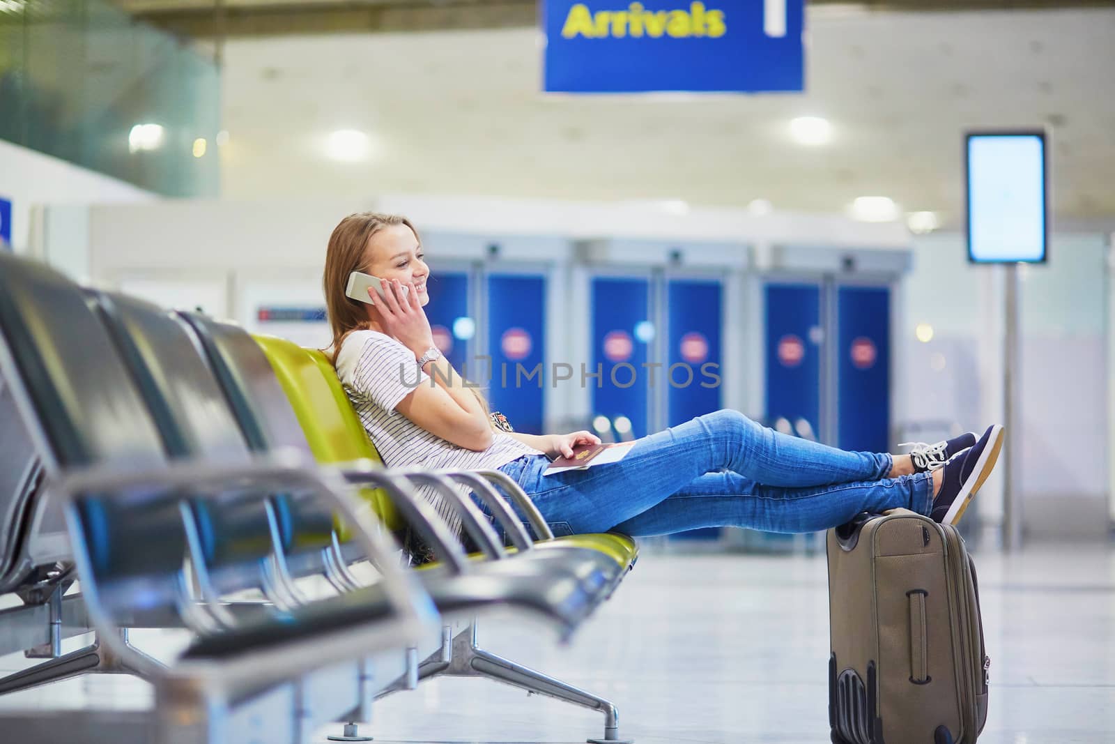 Young traveler with carry on luggage in international airport checking her mobile phone while waiting for her flight