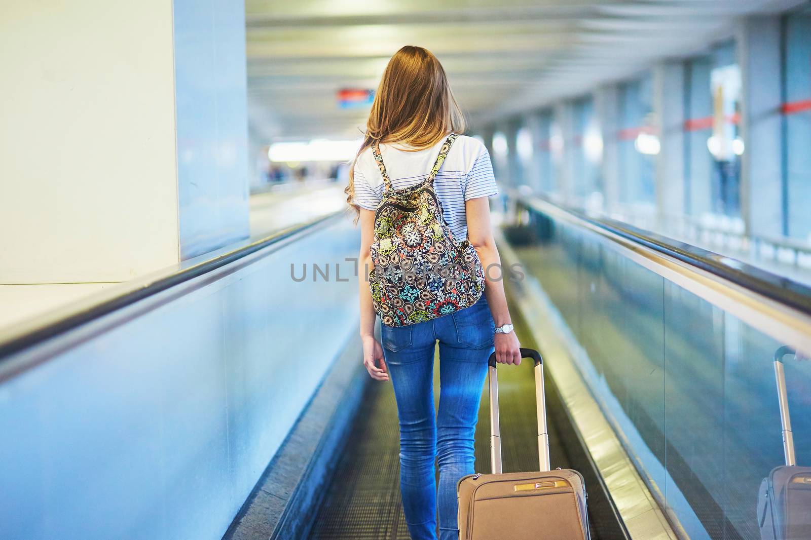 Beautiful young tourist girl with backpack and carry on luggage in international airport, on travelator