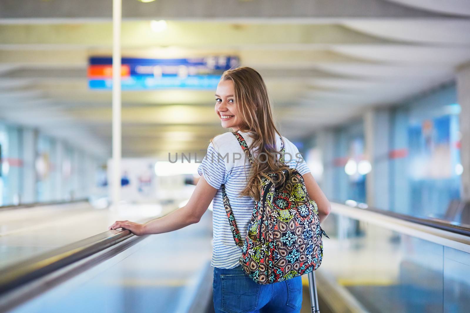 Beautiful young tourist girl with backpack and carry on luggage in international airport, on travelator