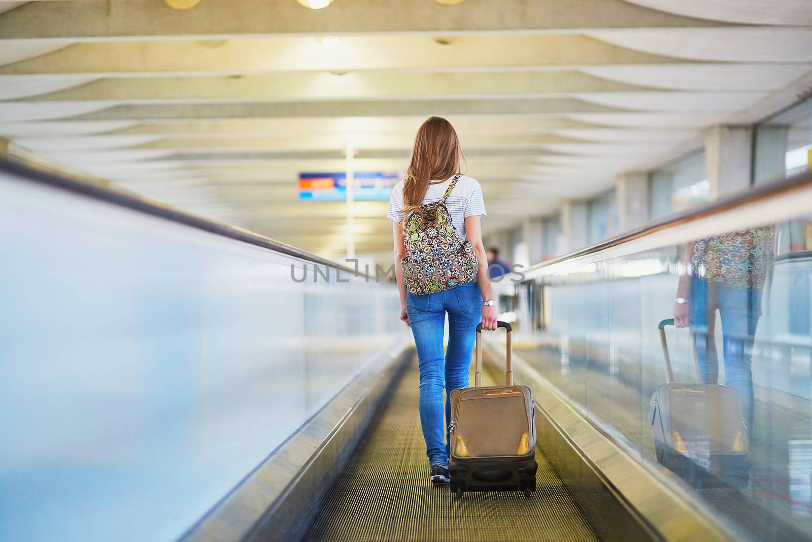 Beautiful young tourist girl with backpack and carry on luggage in international airport, on travelator