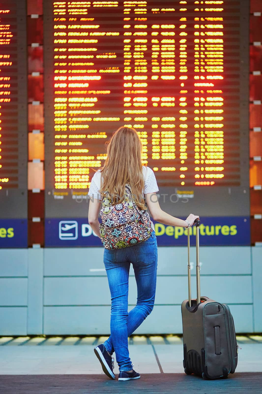 Beautiful young tourist girl with backpack and carry on luggage in international airport, near flight information board