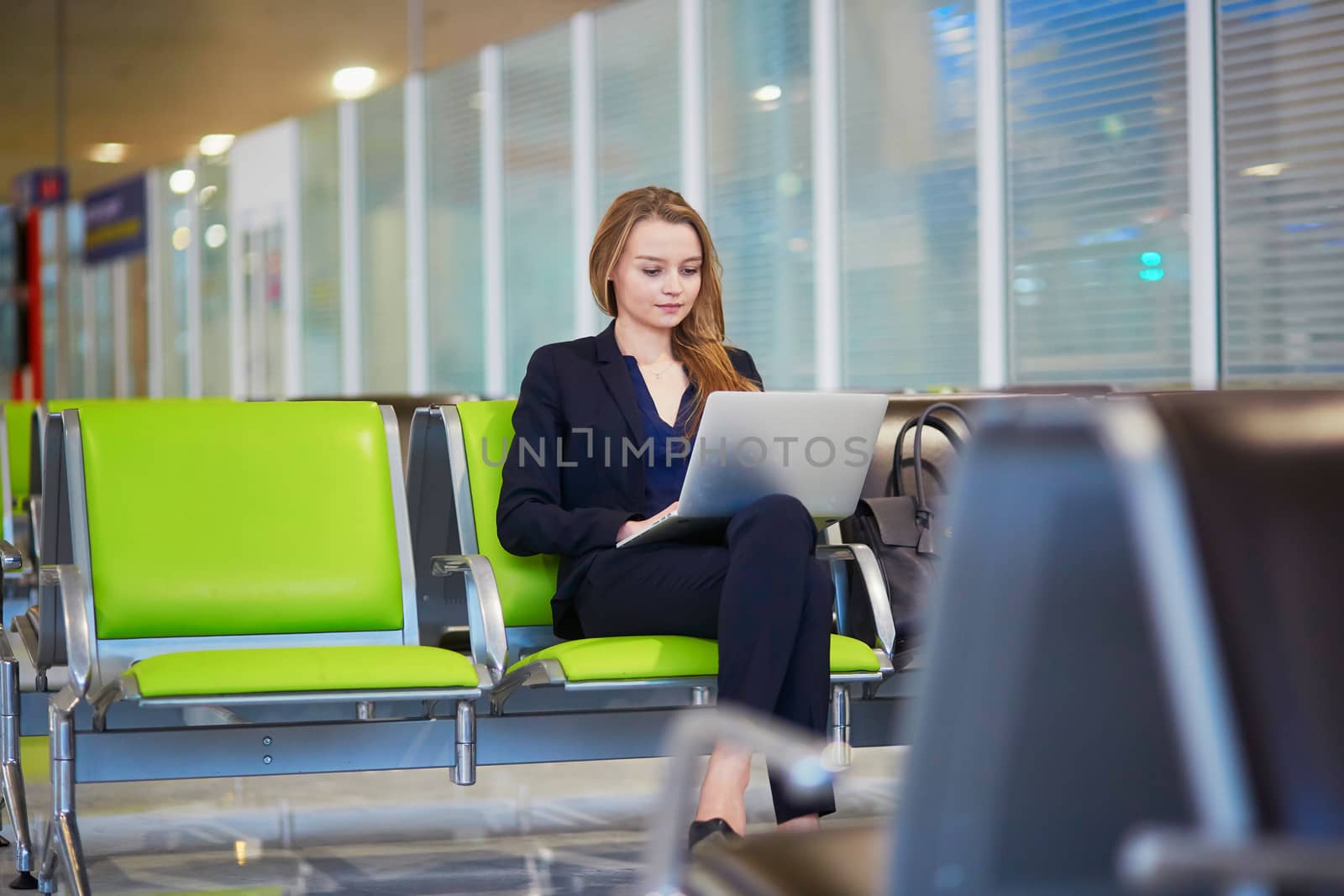 Young elegant business woman with hand luggage in international airport terminal, working on her laptop while waiting for flight