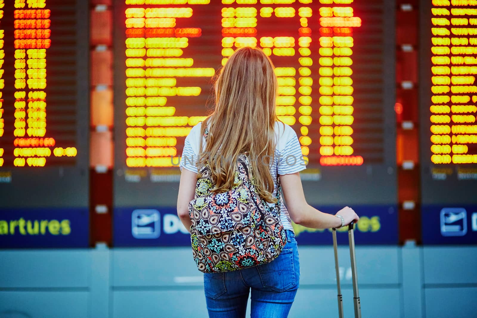 Beautiful young tourist girl with backpack and carry on luggage in international airport, near flight information board