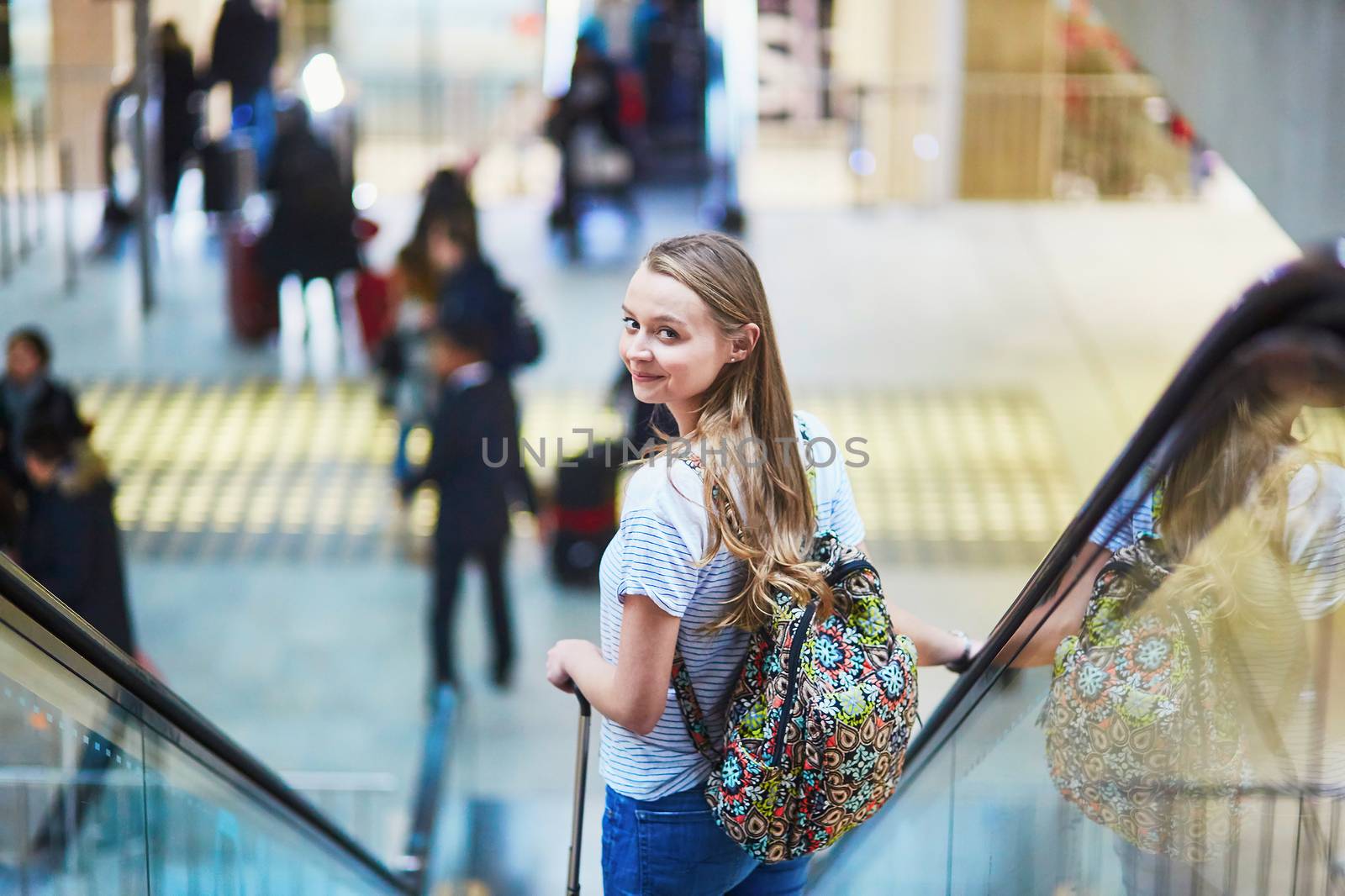 Beautiful young tourist girl with backpack and carry on luggage in international airport, on escalator