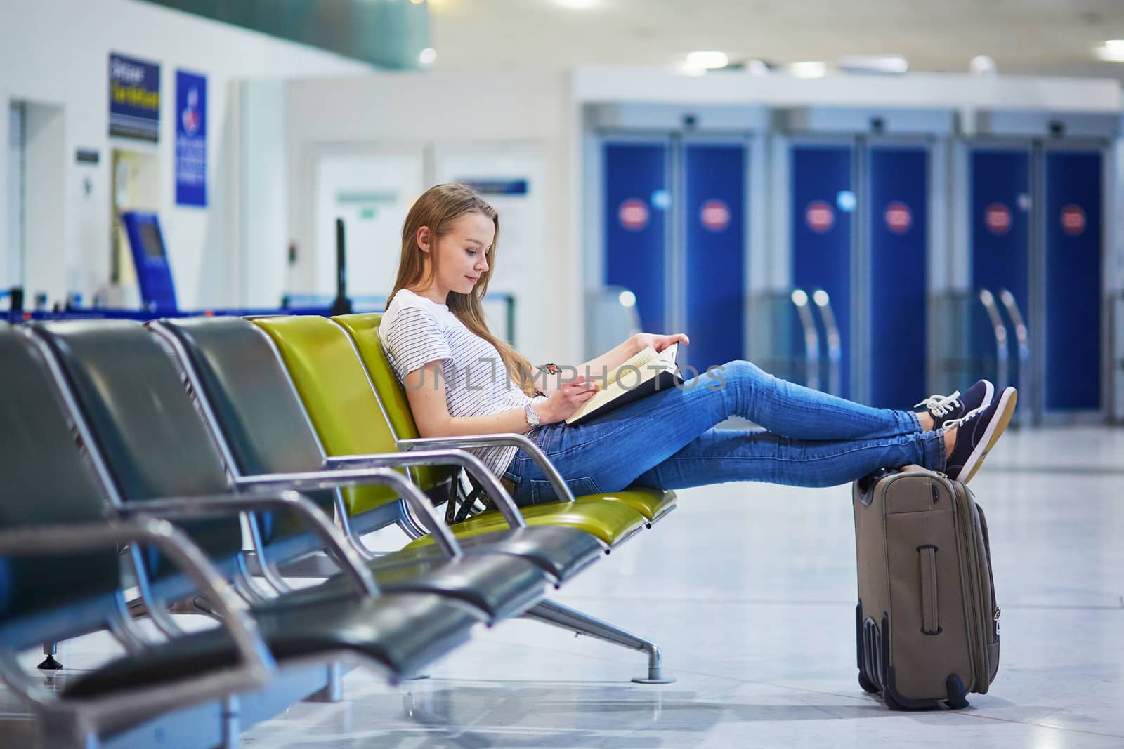 Beautiful young tourist girl with backpack and carry on luggage in international airport, reading a book while waiting for her flight