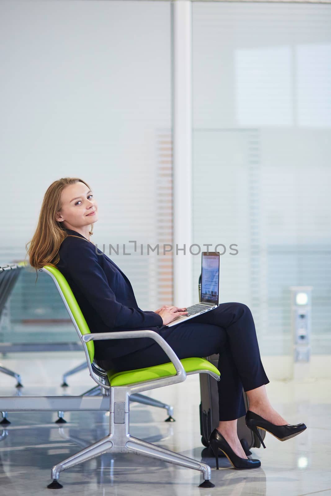 Young elegant business woman with hand luggage in international airport terminal, working on her laptop while waiting for flight