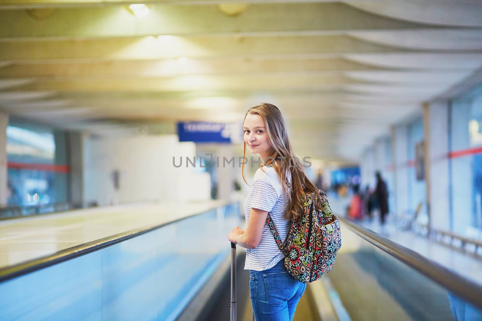 Beautiful young tourist girl with backpack and carry on luggage in international airport, on travelator