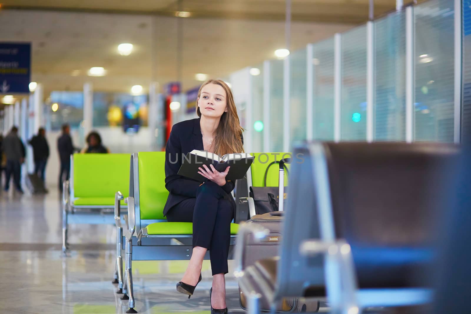 Young elegant business woman with hand luggage in international airport terminal, reading book while waiting for flight