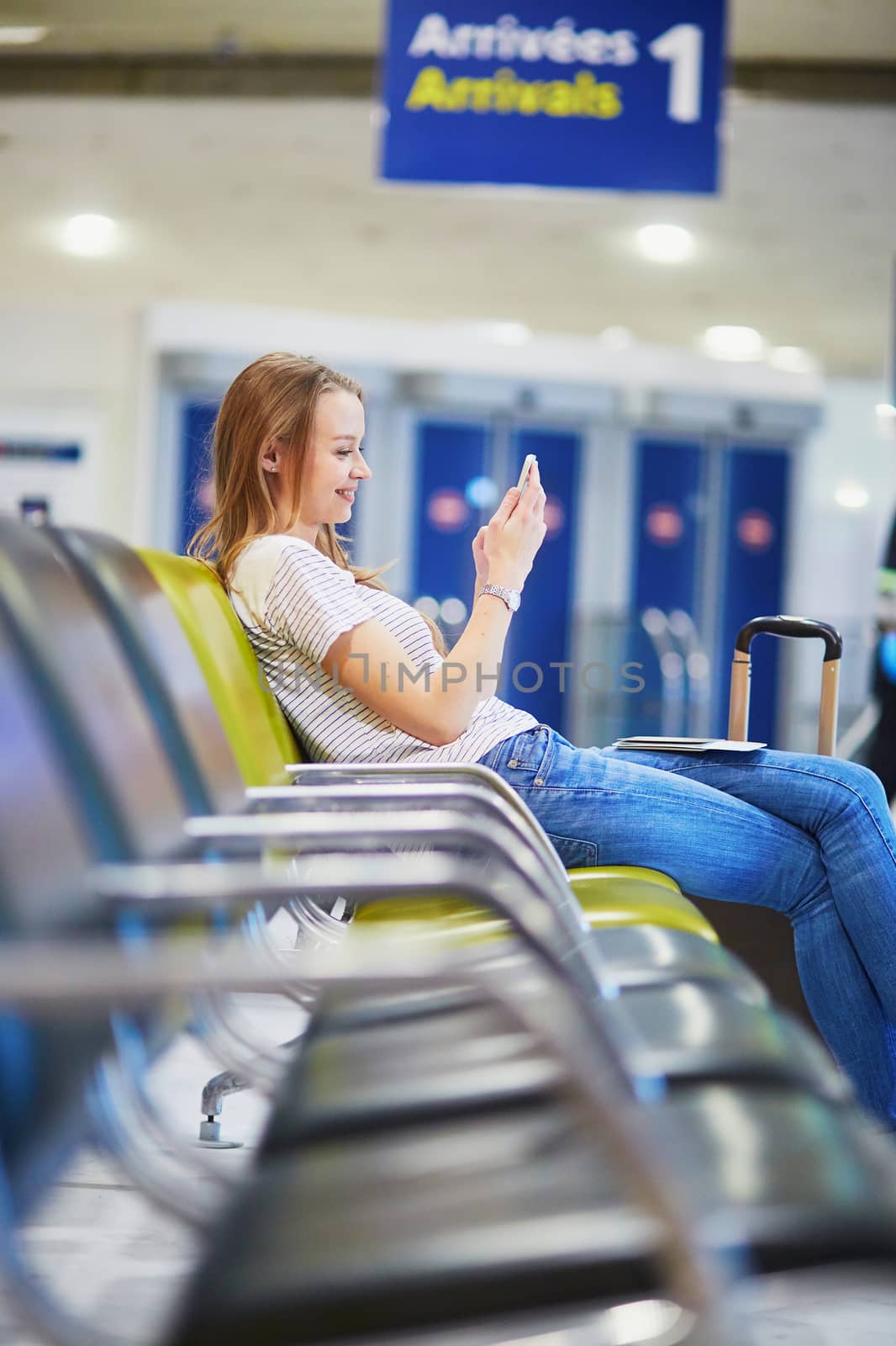 Young traveler with carry on luggage in international airport checking her mobile phone while waiting for her flight