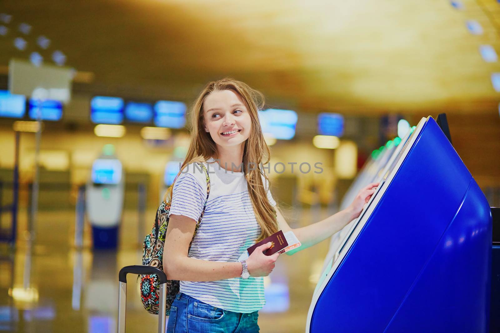 Beautiful young tourist girl with backpack and carry on luggage in international airport, doing self check-in