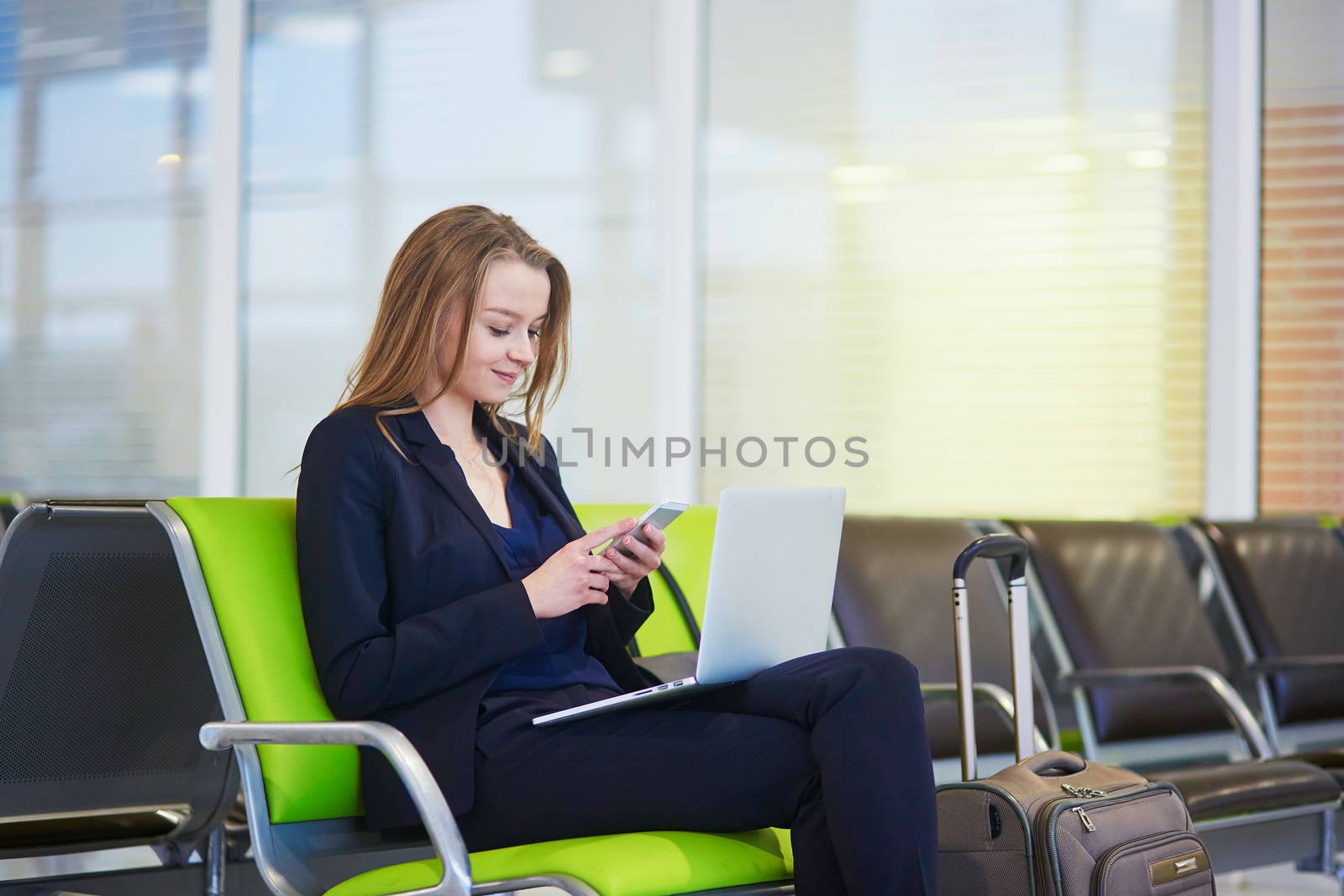 Young elegant business woman with hand luggage in international airport terminal, checking her mobile phone while waiting for flight