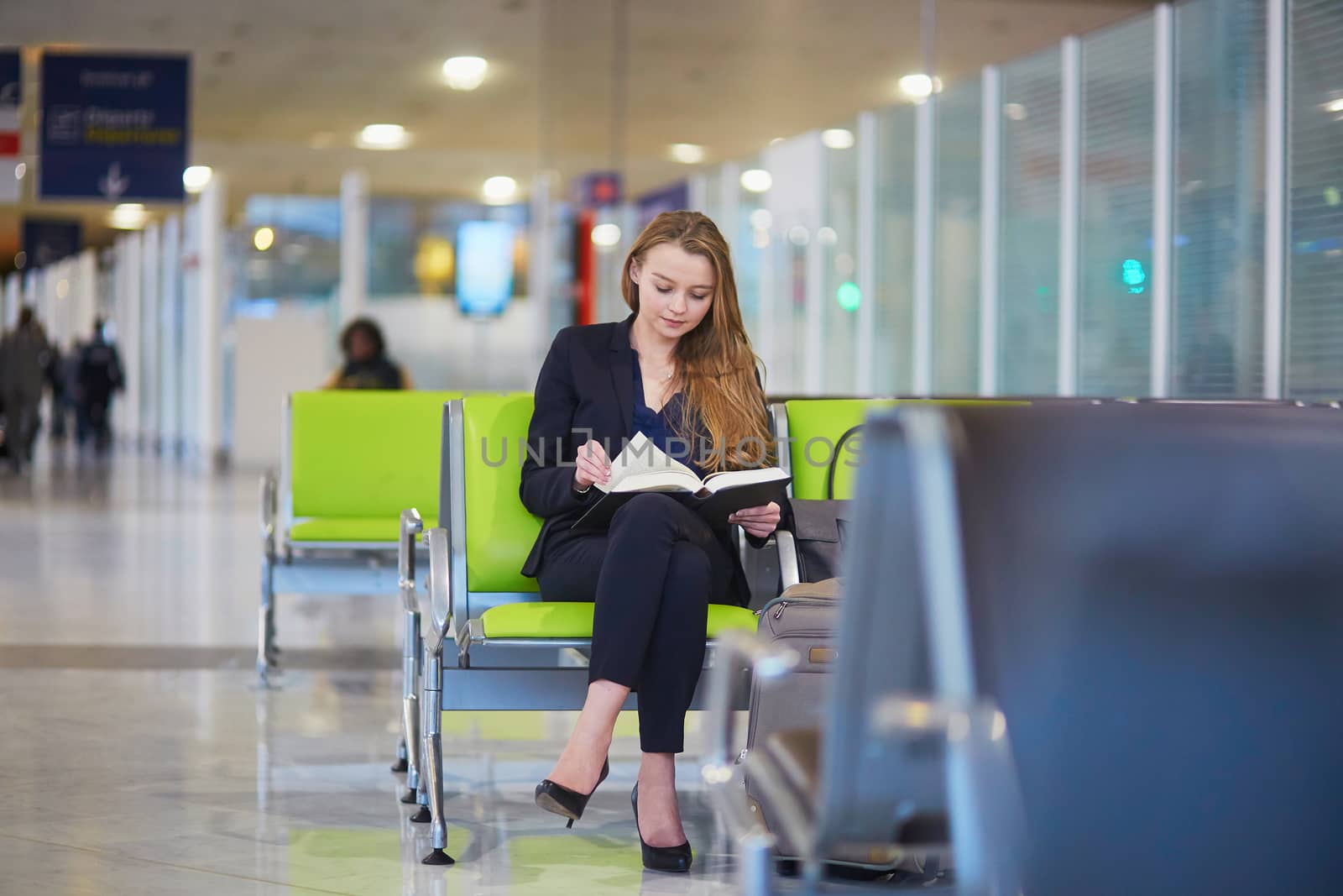 Young elegant business woman with hand luggage in international airport terminal, reading a book while waiting for flight