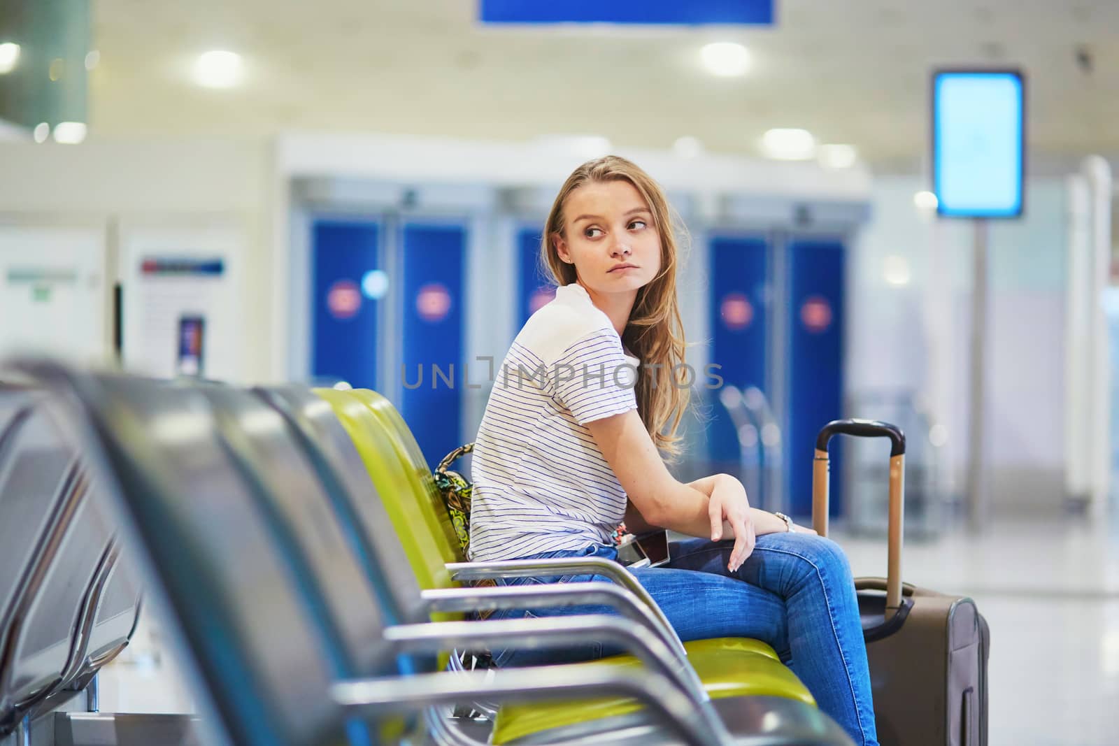 Beautiful young tourist girl with backpack and carry on luggage in international airport, waiting for her flight, looking upset. Delayed or canceled flight concept
