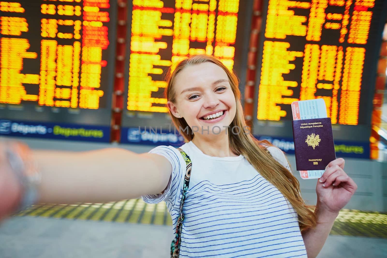 Beautiful young tourist girl in international airport, taking funny selfie with passport and boarding pass near flight information board