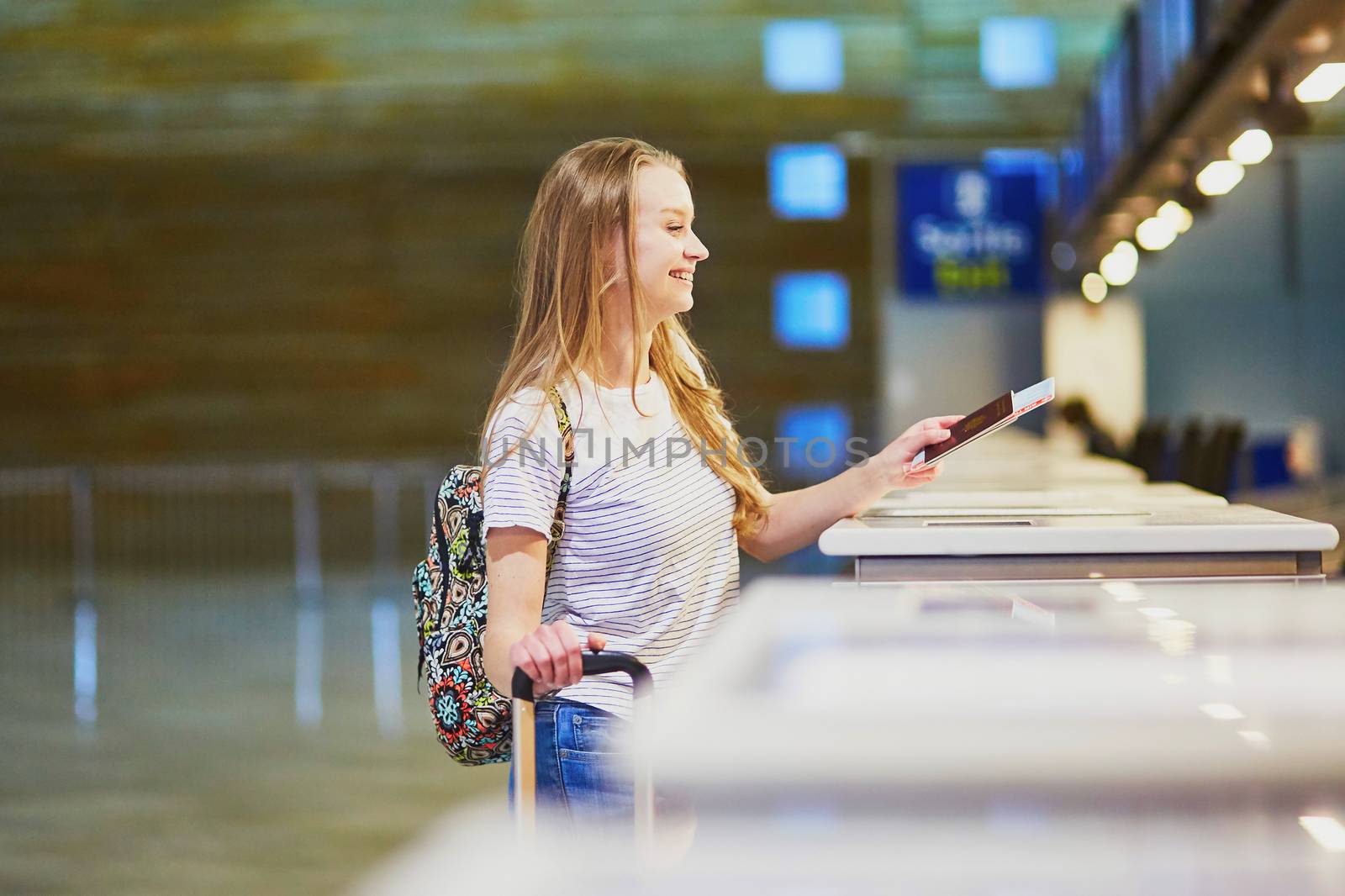 Beautiful young tourist girl with backpack and carry on luggage in international airport at check-in counter, giving her passport to an officer and waiting for her boarding pass