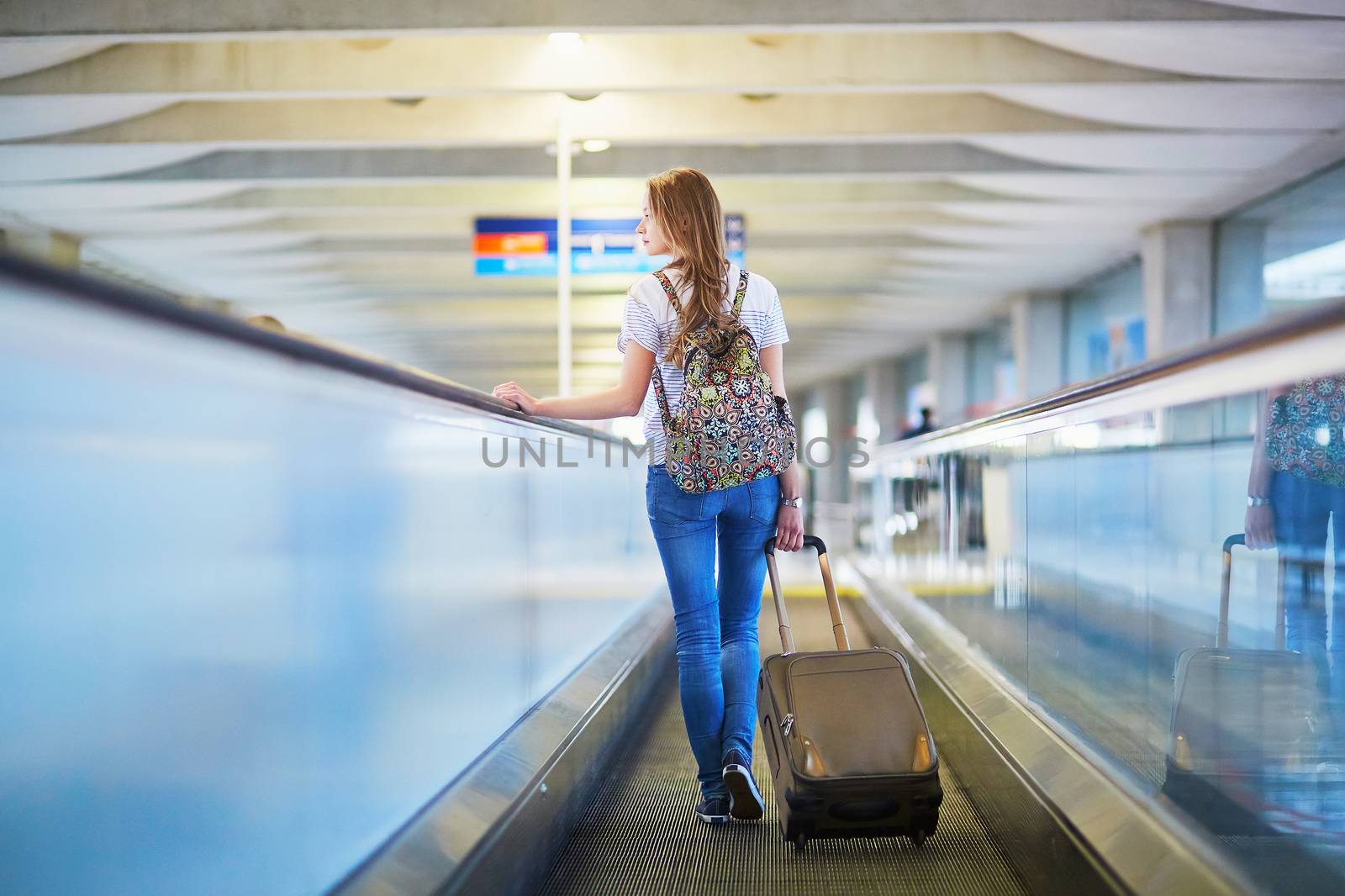 Beautiful young tourist girl with backpack and carry on luggage in international airport, on travelator