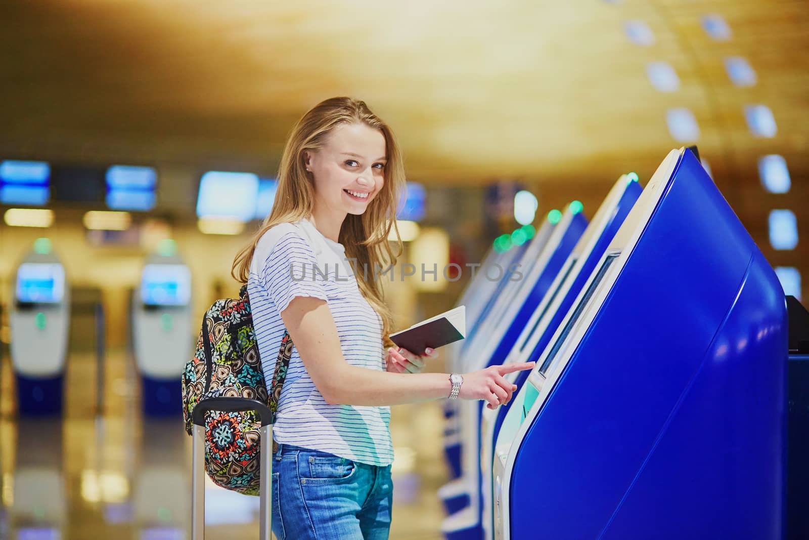 Beautiful young tourist girl with backpack and carry on luggage in international airport, doing self check-in