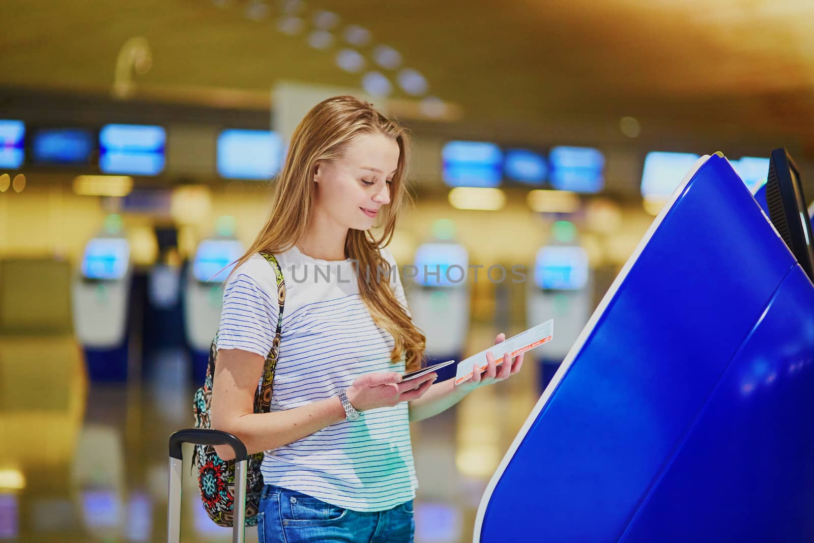 Beautiful young tourist girl with backpack and carry on luggage in international airport, doing self check-in