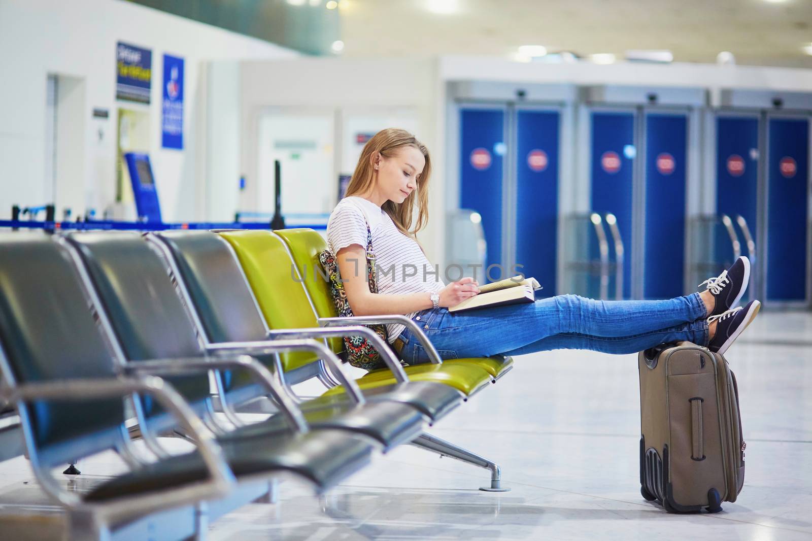 Young traveler with carry on luggage in international airport reading a book while waiting for her flight