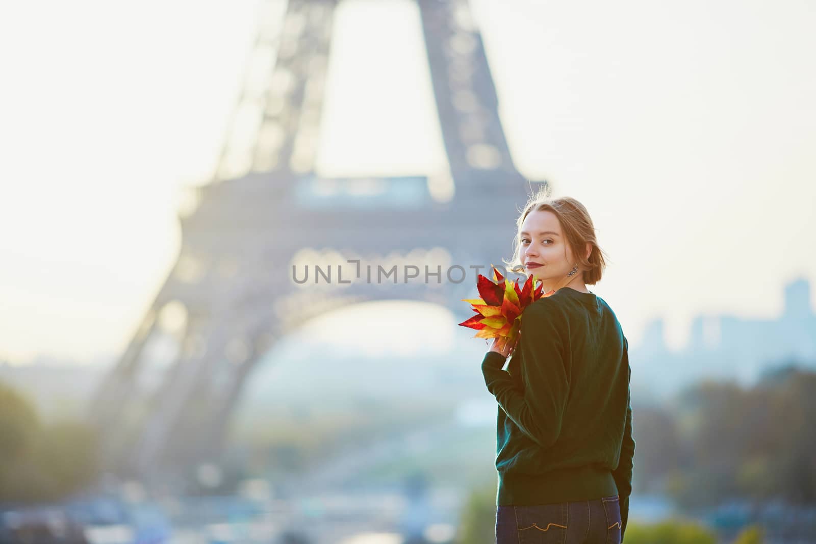 Beautiful young French woman with bunch of colorful autumn leaves near the Eiffel tower in Paris on a fall day