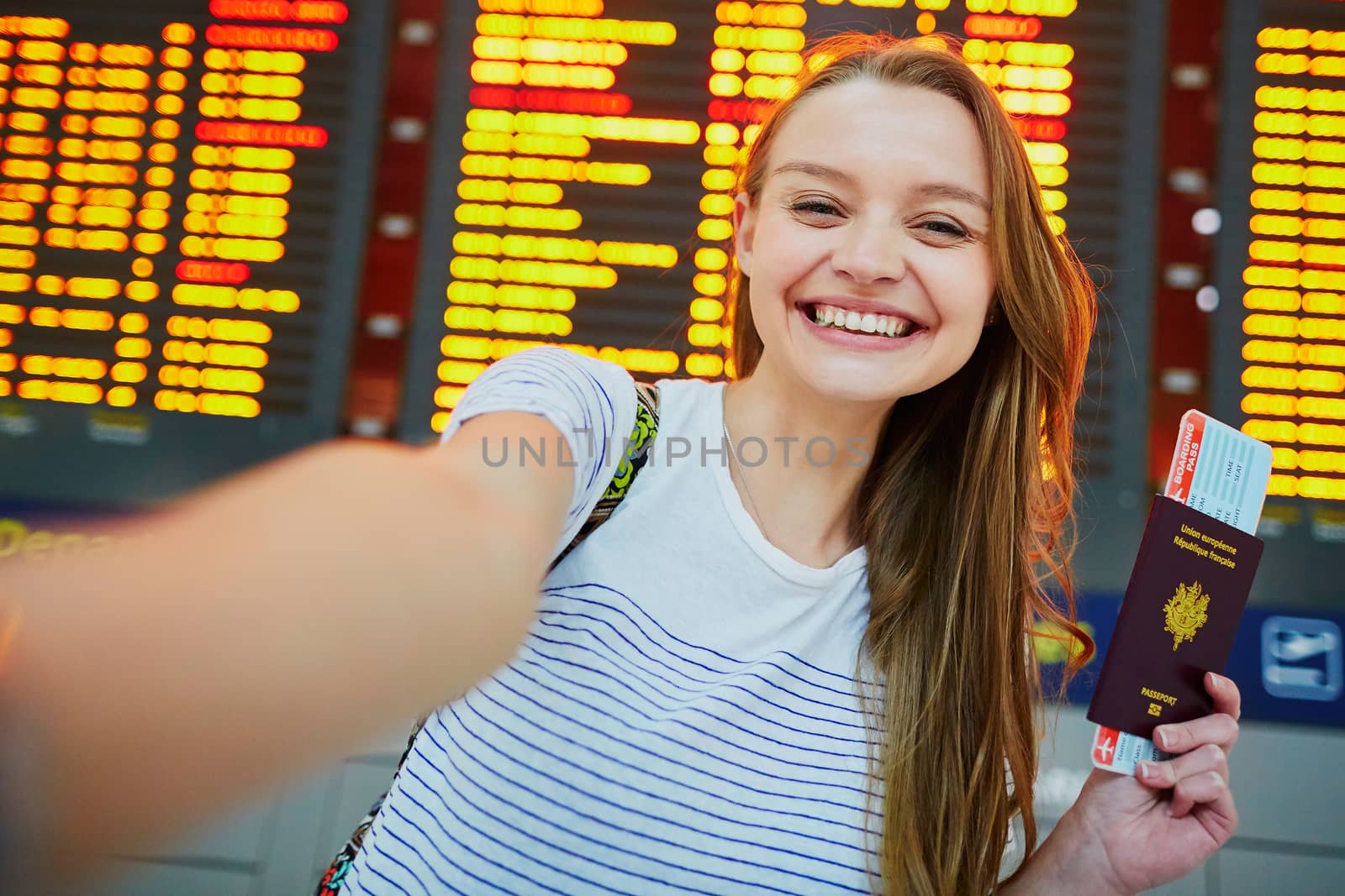 Beautiful young tourist girl in international airport, taking funny selfie with passport and boarding pass near flight information board