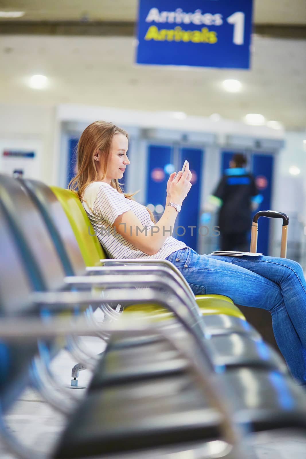 Young traveler with carry on luggage in international airport checking her mobile phone while waiting for her flight