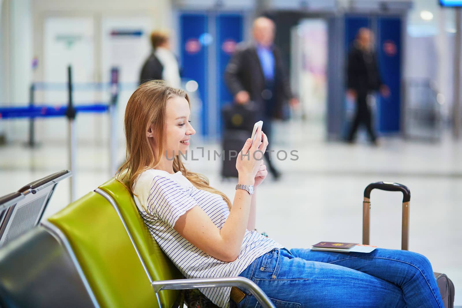Young traveler with carry on luggage in international airport checking her mobile phone while waiting for her flight