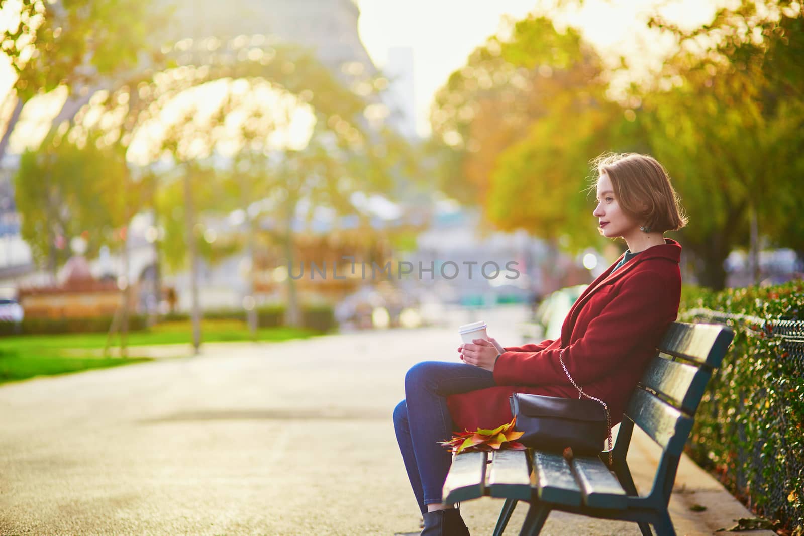 Beautiful young French woman with bunch of colorful autumn leaves sitting on the bench near the Eiffel tower in Paris on a fall day