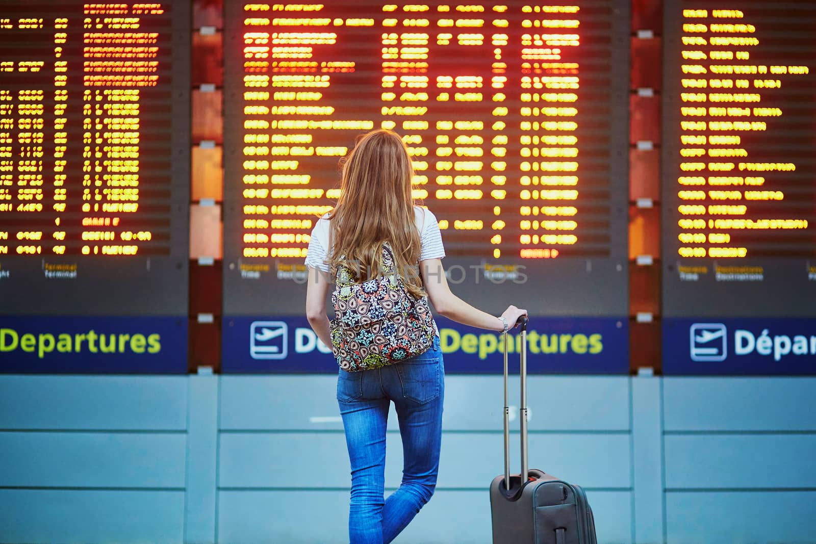 Beautiful young tourist girl with backpack and carry on luggage in international airport, near flight information board