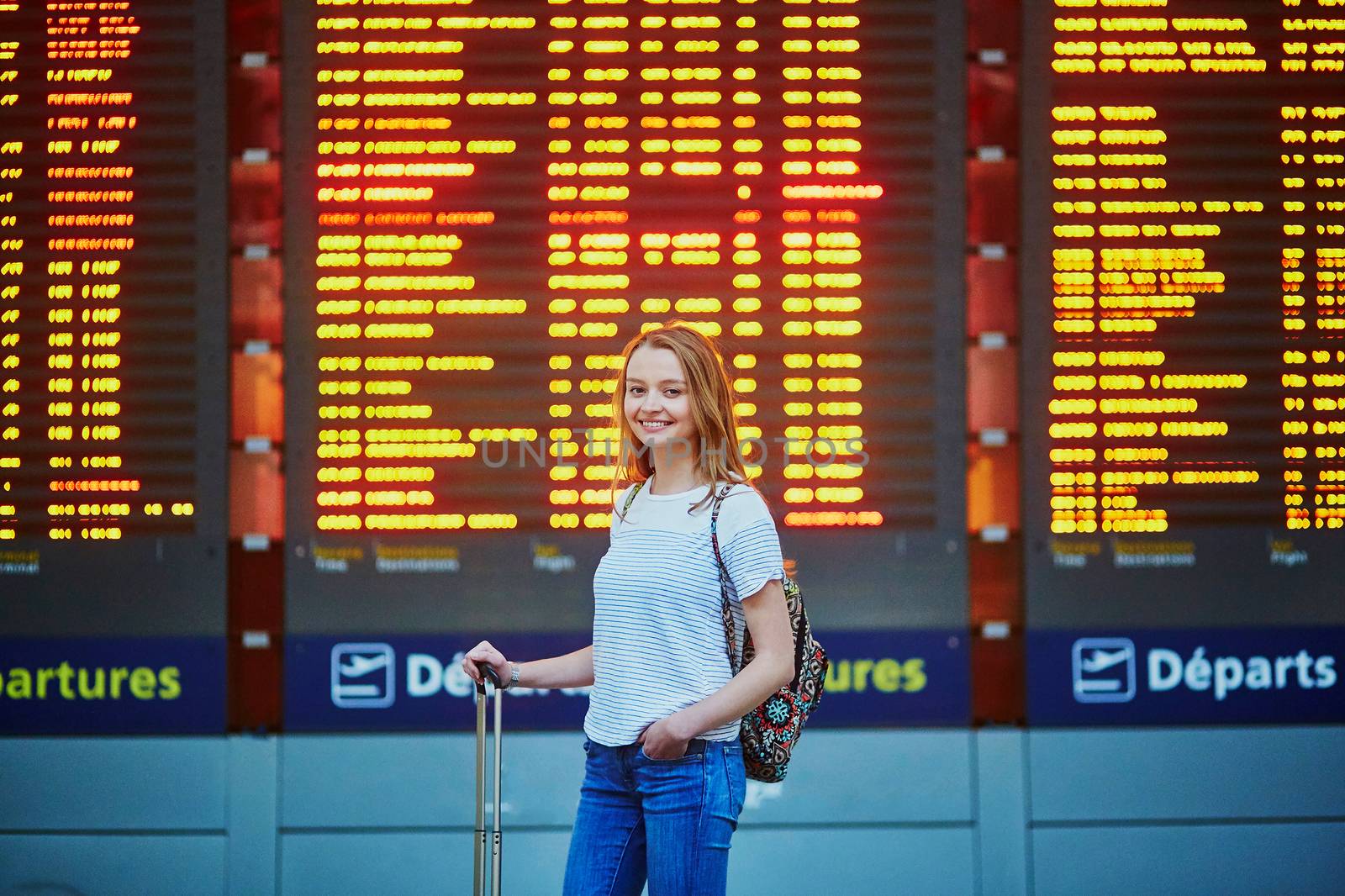 Beautiful young tourist girl with backpack and carry on luggage in international airport, near flight information board