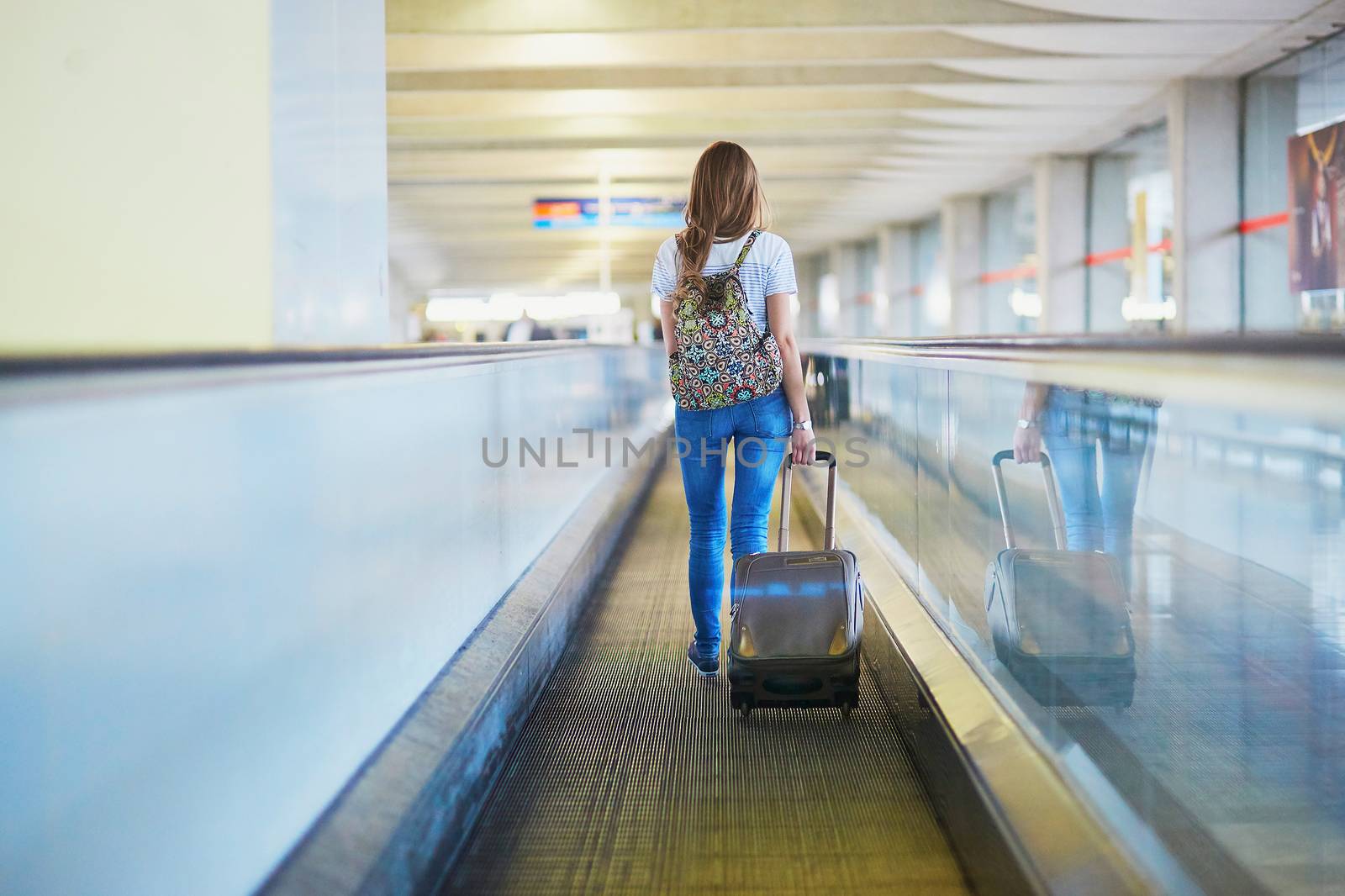 Beautiful young tourist girl with backpack and carry on luggage in international airport, on travelator