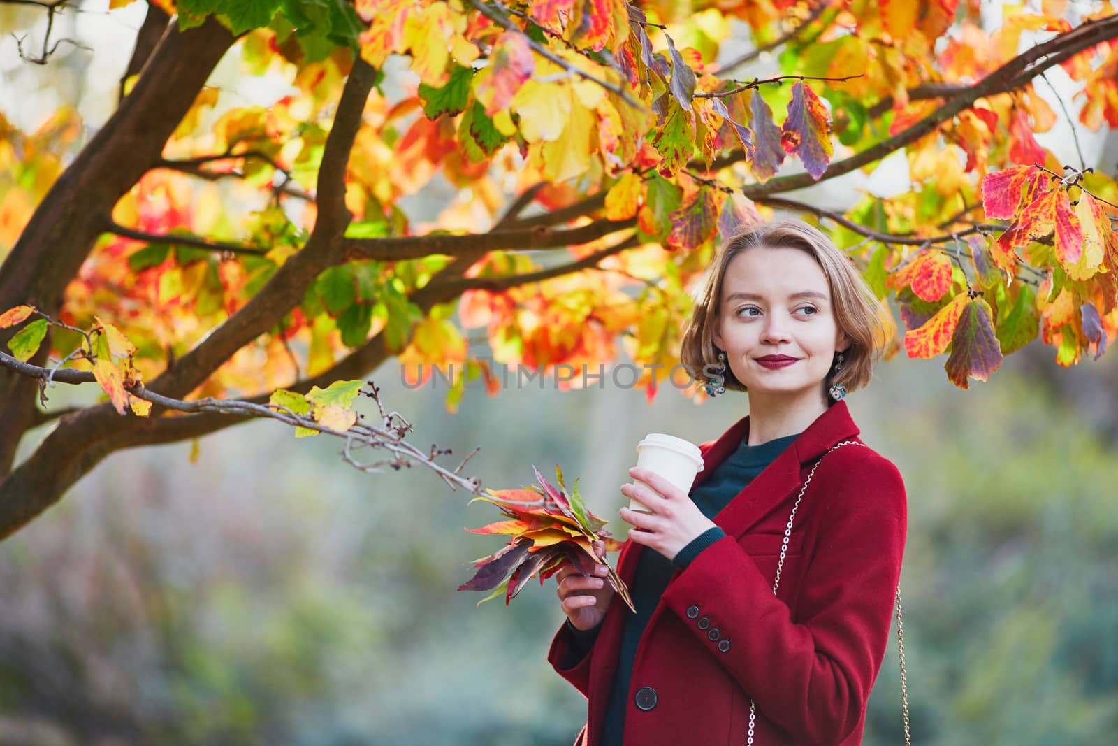 Beautiful young woman with bunch of colorful autumn leaves walking in park on a fall day and drinking coffee to go