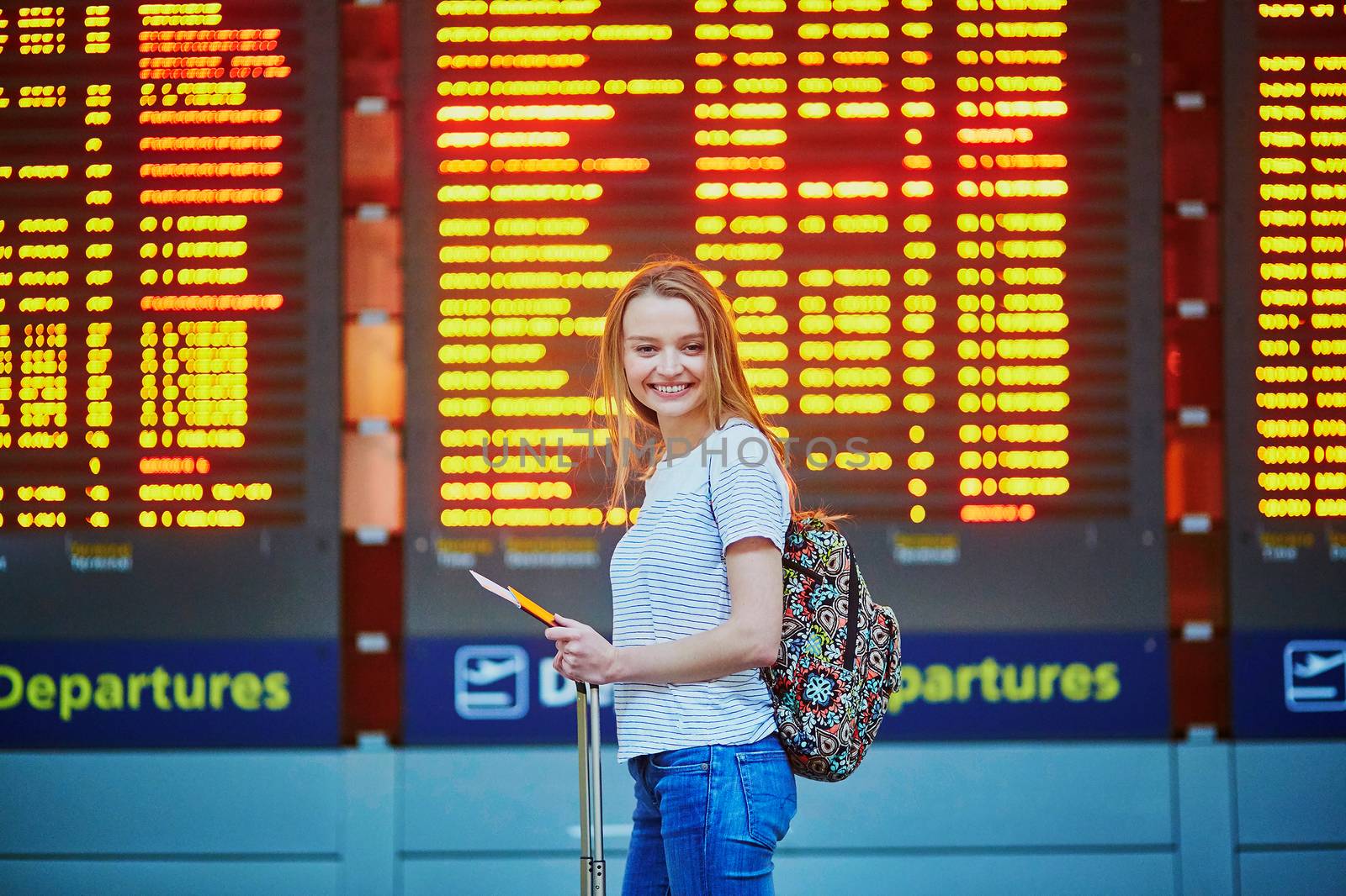 Beautiful young tourist girl with backpack and carry on luggage in international airport, near flight information board